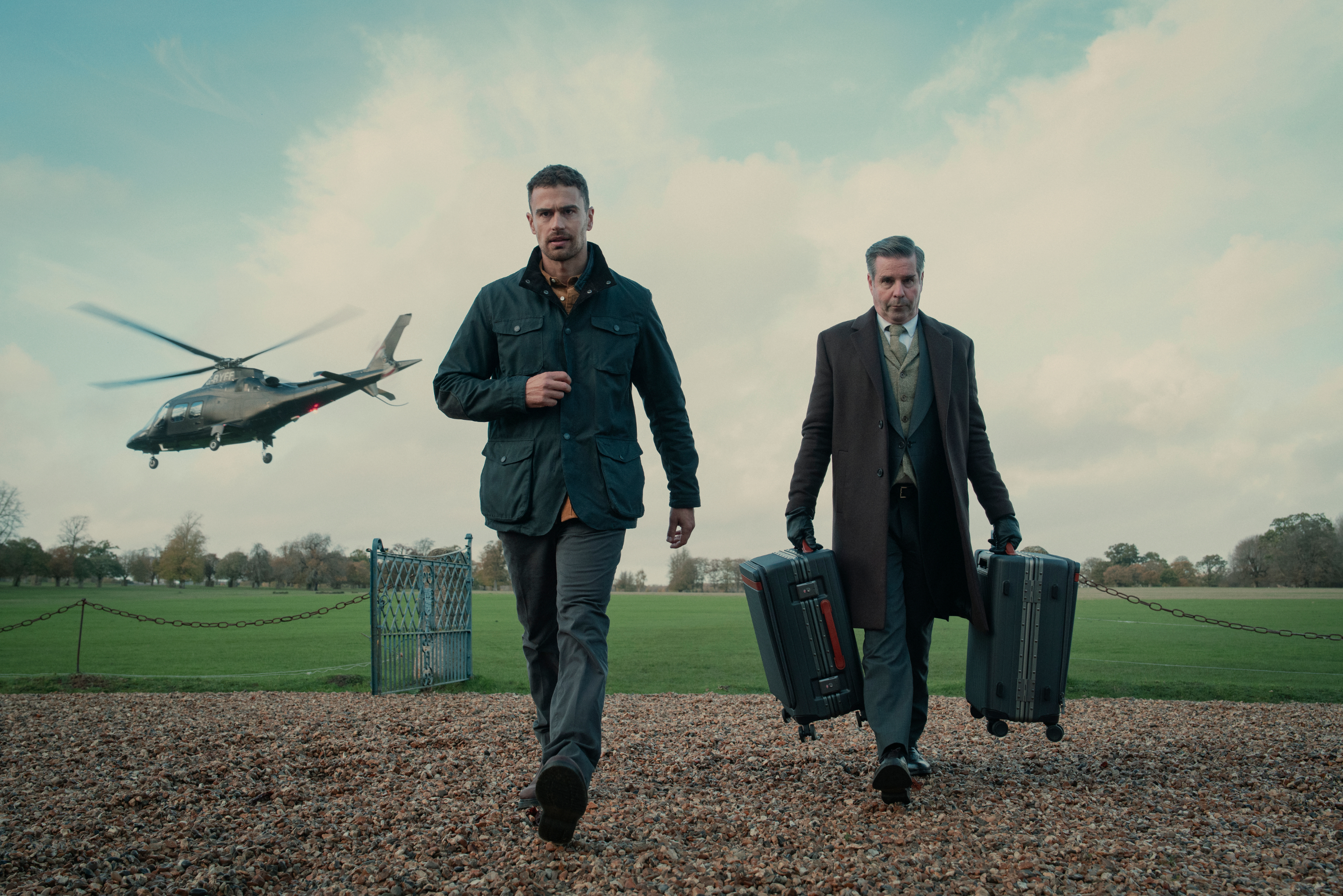 two men in jackets walking away from a helicopter, one has a luggage roller in each hand