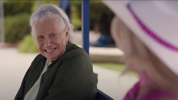 An older woman sitting on a bench, smiliing, as seen in "barbie"