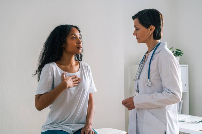 Mature healthcare worker showing concern about female African-American patient who is touching her chest and describing painful ache to cardiac doctor in hospital office