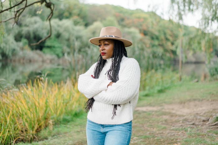 Woman in a white sweater and jeans with arms crossed wearing a wide-brimmed hat, outdoors