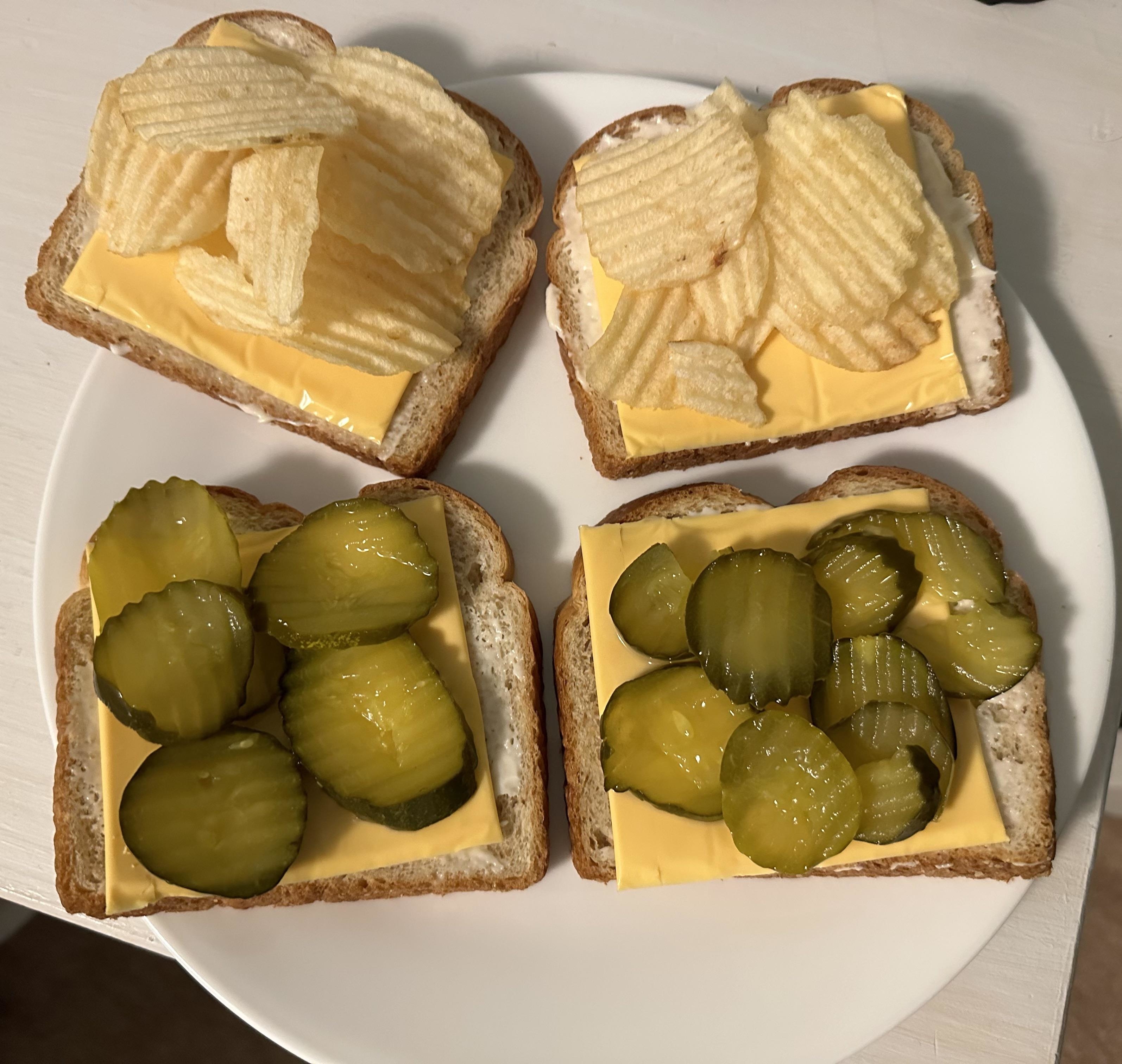 Four slices of white bread with pickles and American cheese slices on half and potato chips and more cheese on the other half