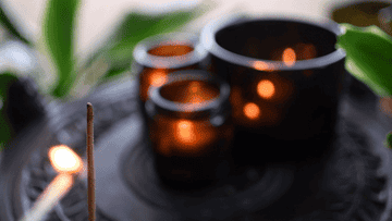 Lit matchstick near three small candles emitting a soft glow on a dark tray surrounded by greenery
