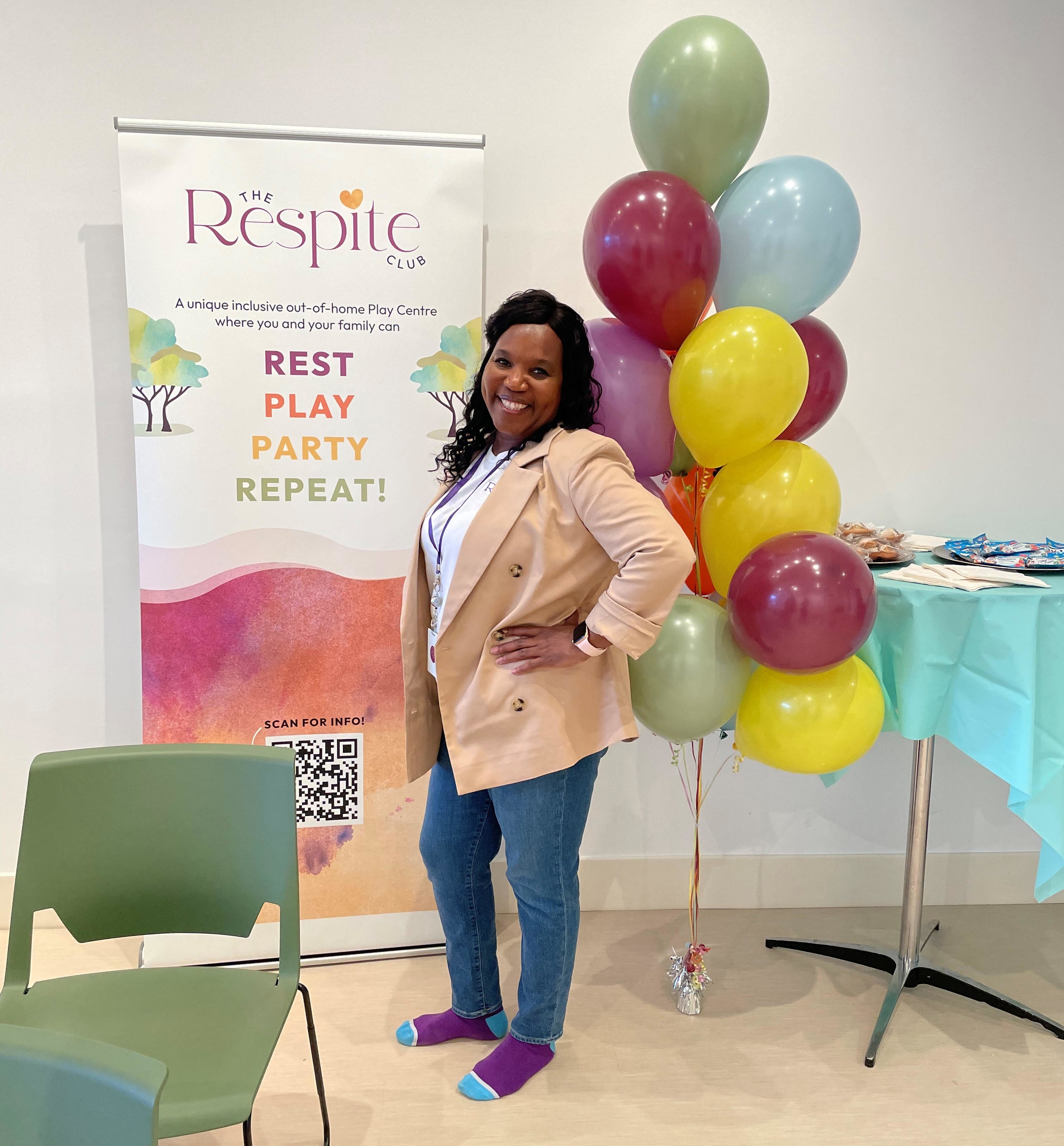Woman posing with hand on hip, smiling, standing by balloons and a sign for The Respite Club. She wears a blazer and jeans