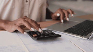 Person using a calculator next to a laptop on a desk, paperwork beneath hands