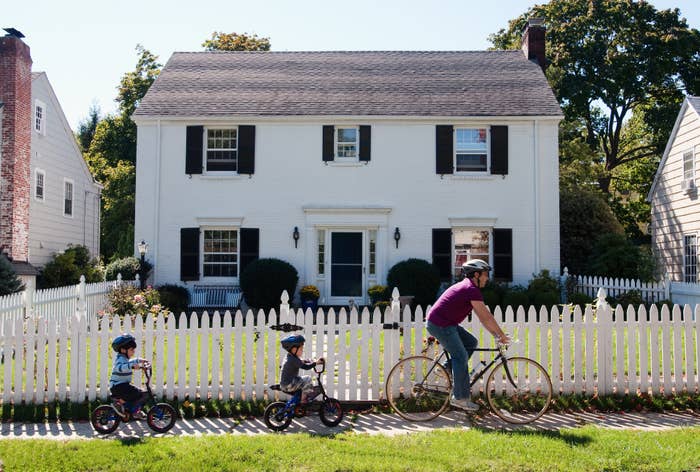 Dad and two young kids riding bikes past a house with a white picket fence