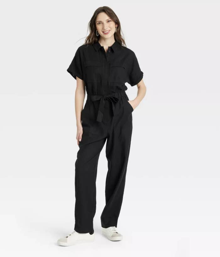 model in a black jumpsuit with short sleeves and a waist tie, paired with white sneakers