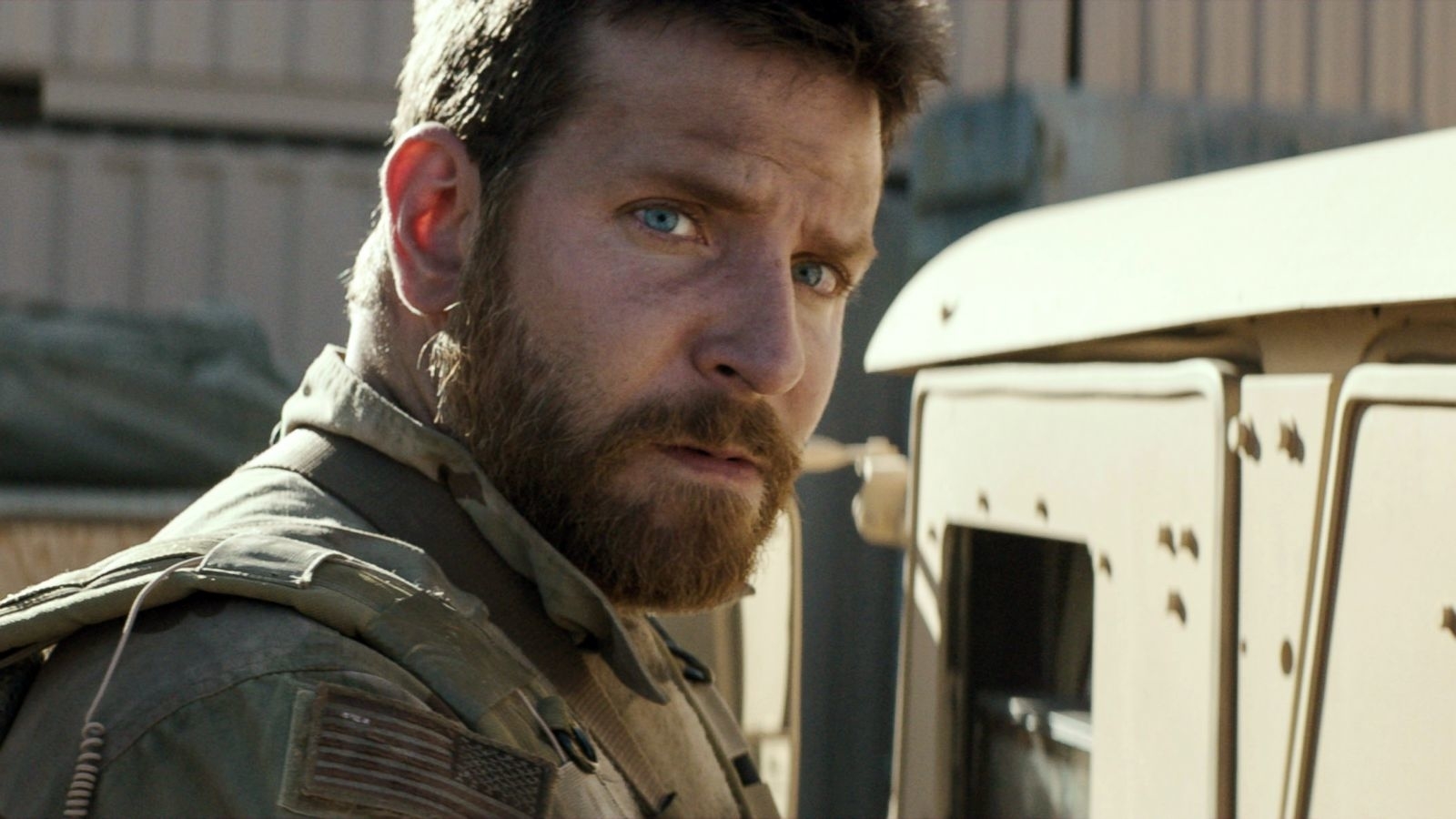 Bearded man in military attire, intense gaze, standing near a vehicle