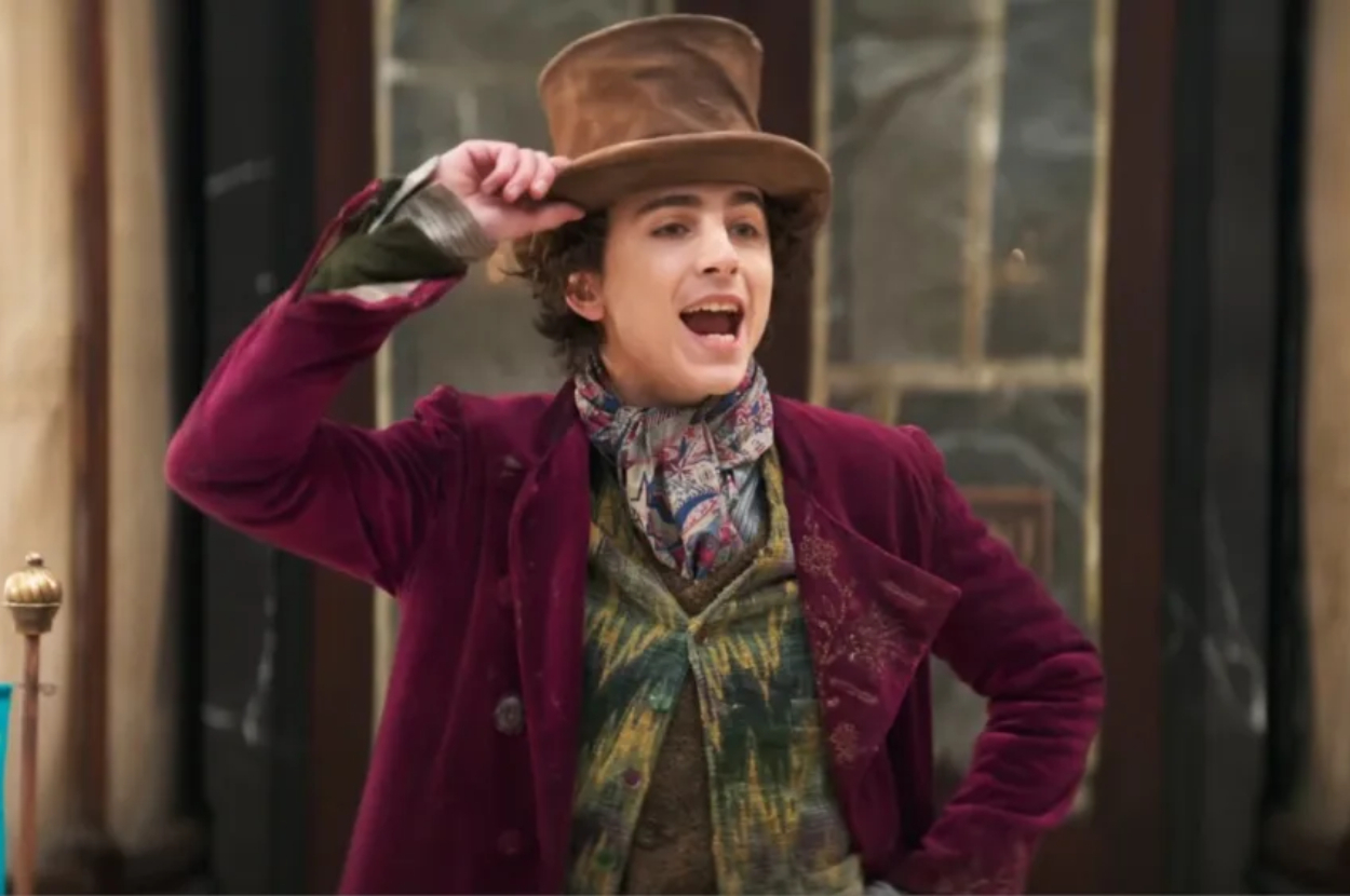A character dressed as Willy Wonka tips his hat while smiling in a scene from a production
