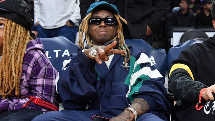Lil Wayne sitting courtside at a basketball game, sporting a navy tracksuit and throwing a peace sign