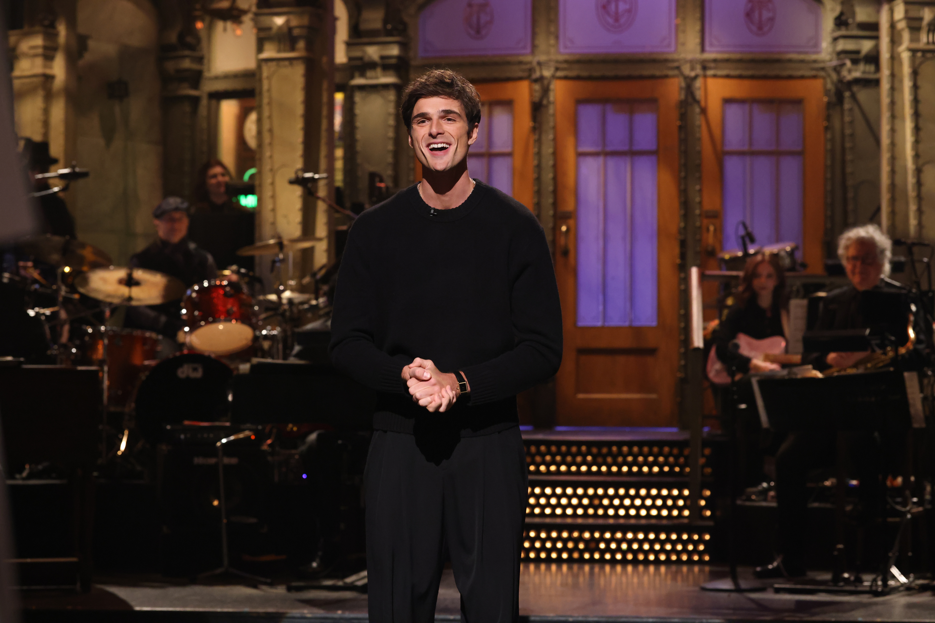 Jacob onstage at SNL with a microphone stand, smiling, wearing a sweater, with the SNL band in the background