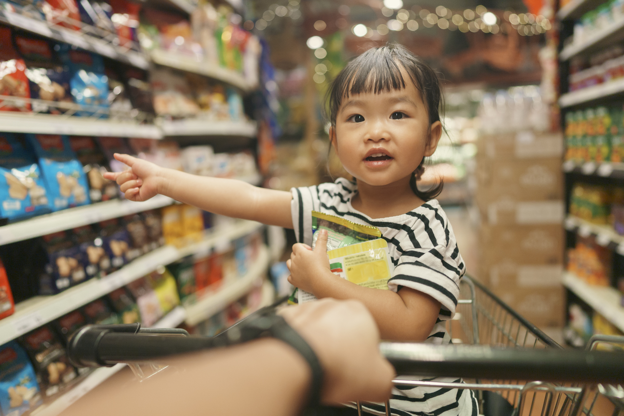 Toddler in a shopping cart pointing at grocery store shelves, appearing curious