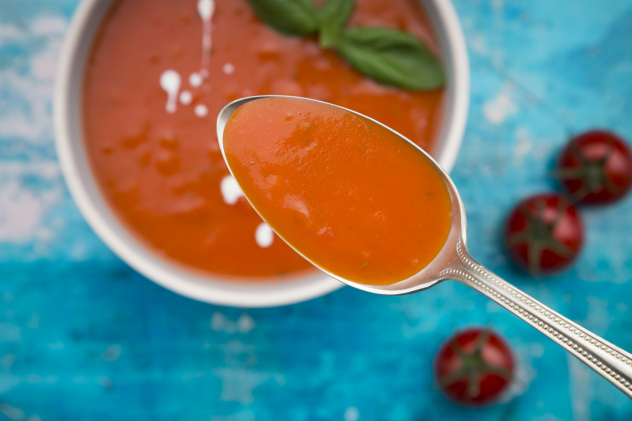 A spoonful of tomato soup above a bowl, with basil leaves and tomatoes nearby