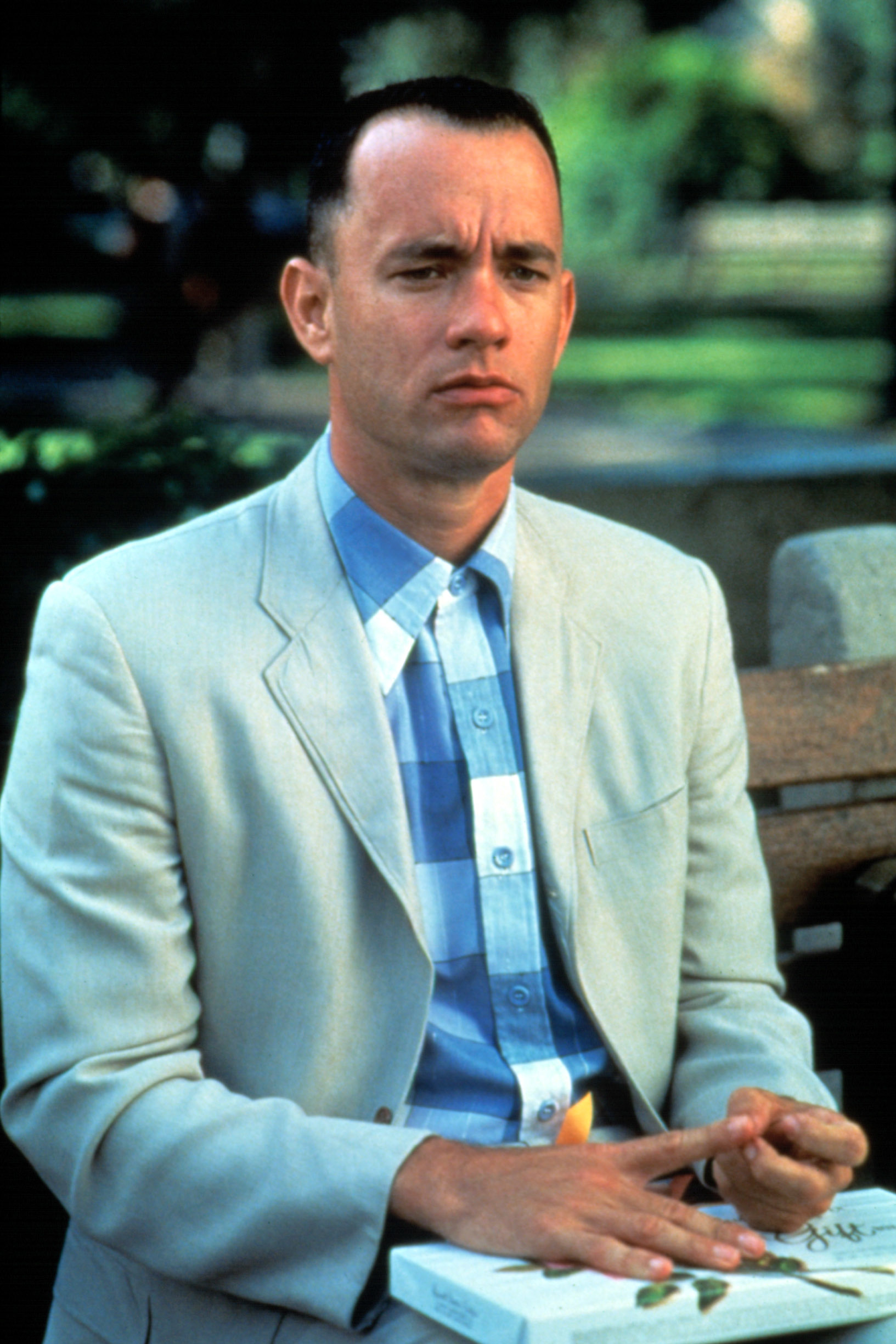 Tom Hanks as Forrest Gump, seated on a bench, wearing a light suit and plaid shirt, holding a briefcase