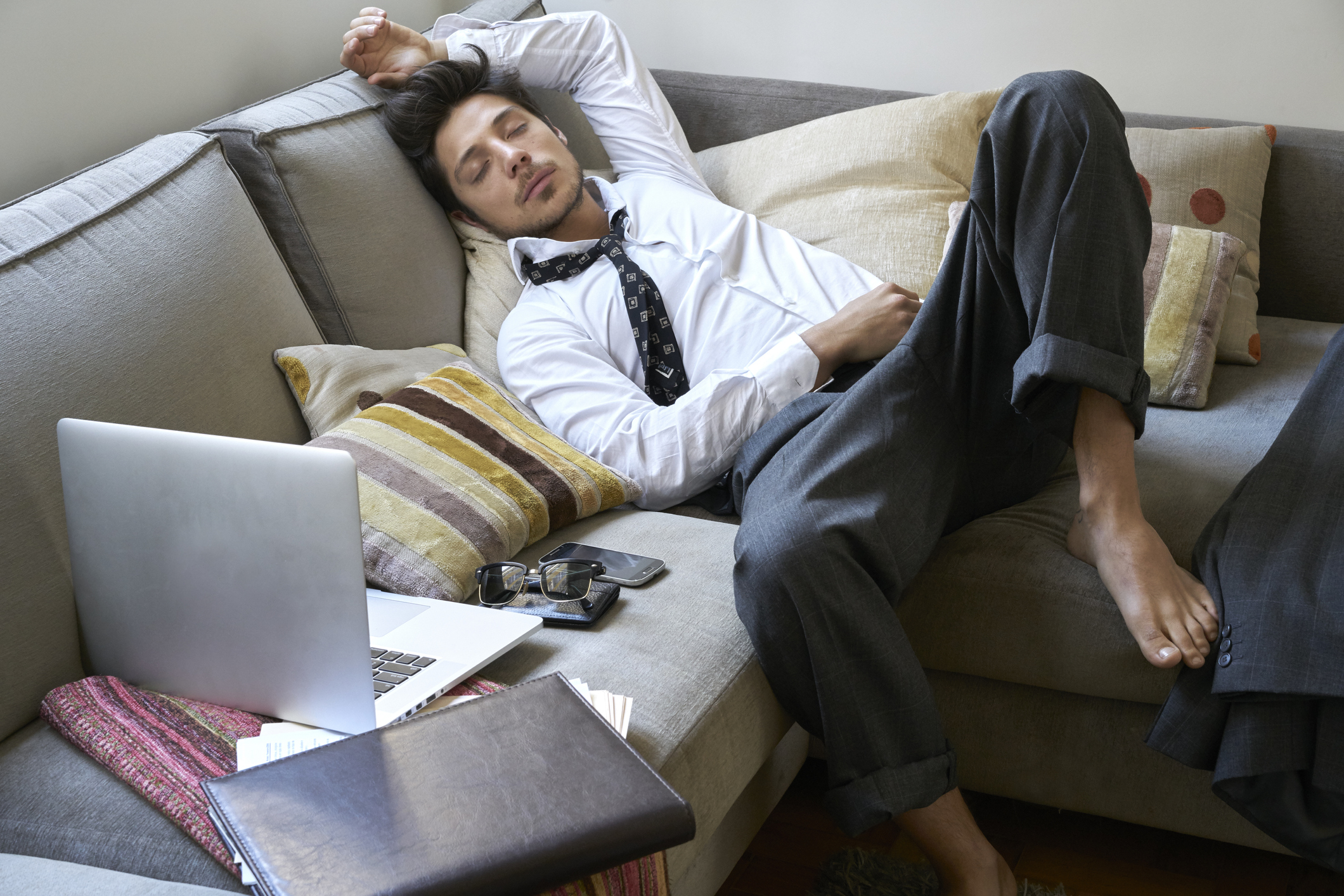 Person in business attire napping on couch with laptop and notebook nearby