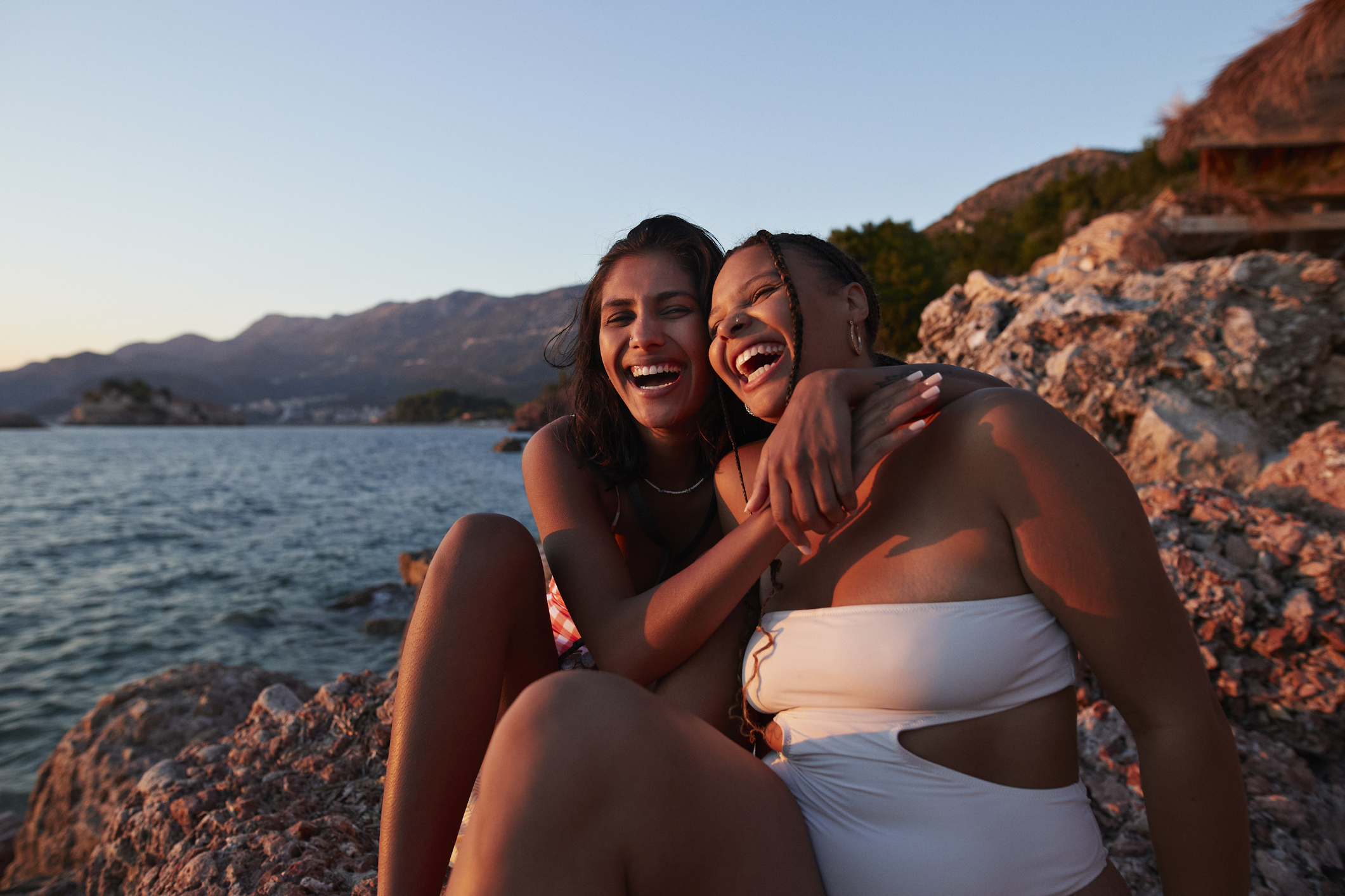 Two people laughing and sitting close together by the water at sunset