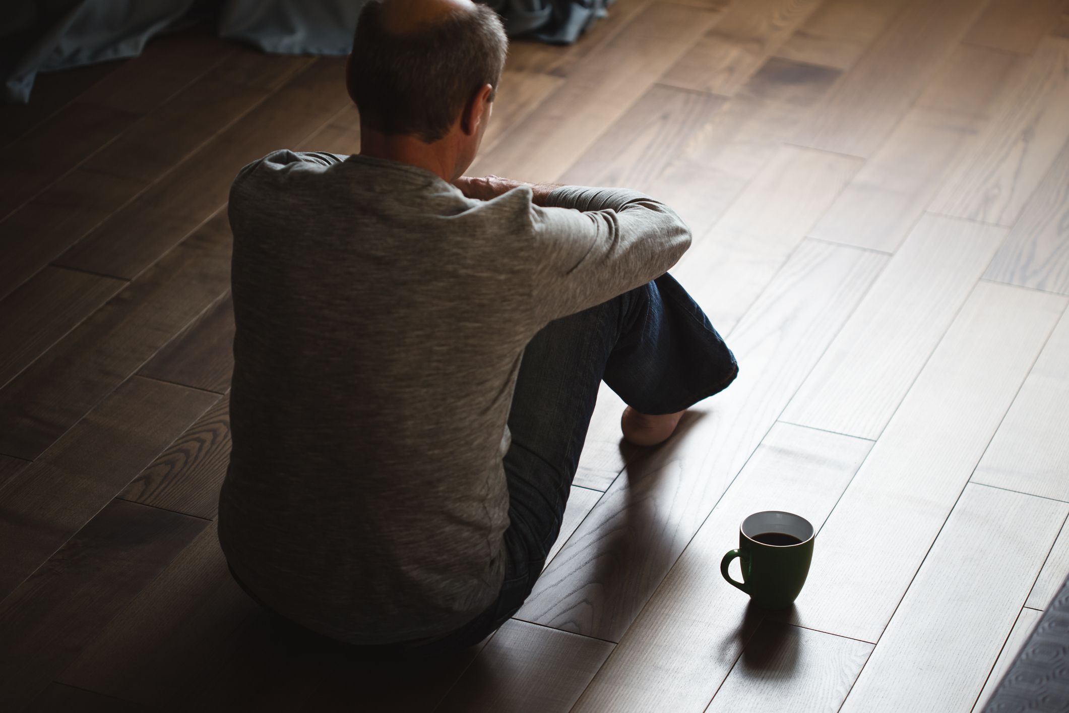 A person deep in thought, sitting on floor with back to camera, holding knees, near a mug