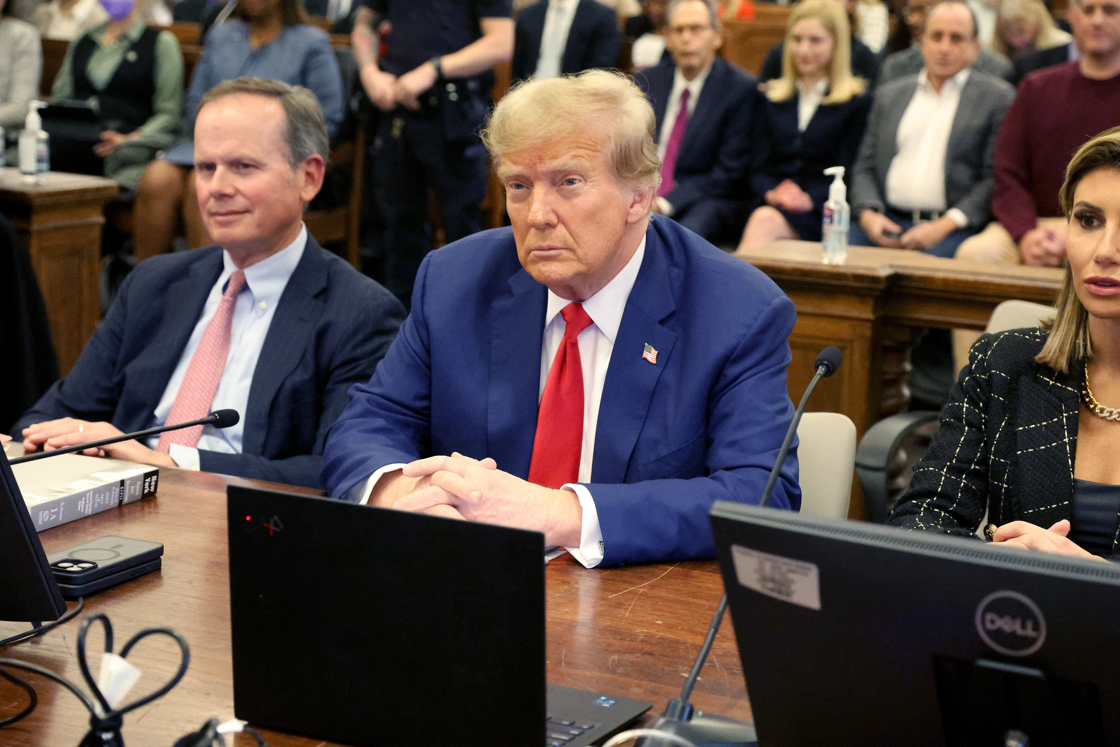 Three people seated at a table, the middle person is Donald Trump. There are onlookers in the background
