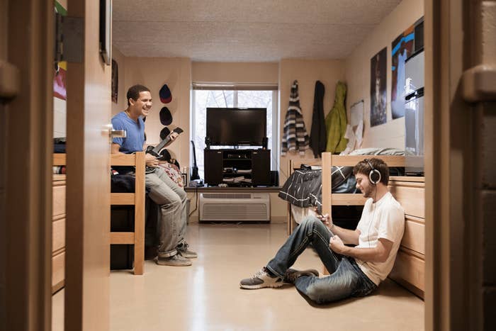 Two people in a dorm room, one seated at a desk with a computer, the other on the floor with headphones and a laptop