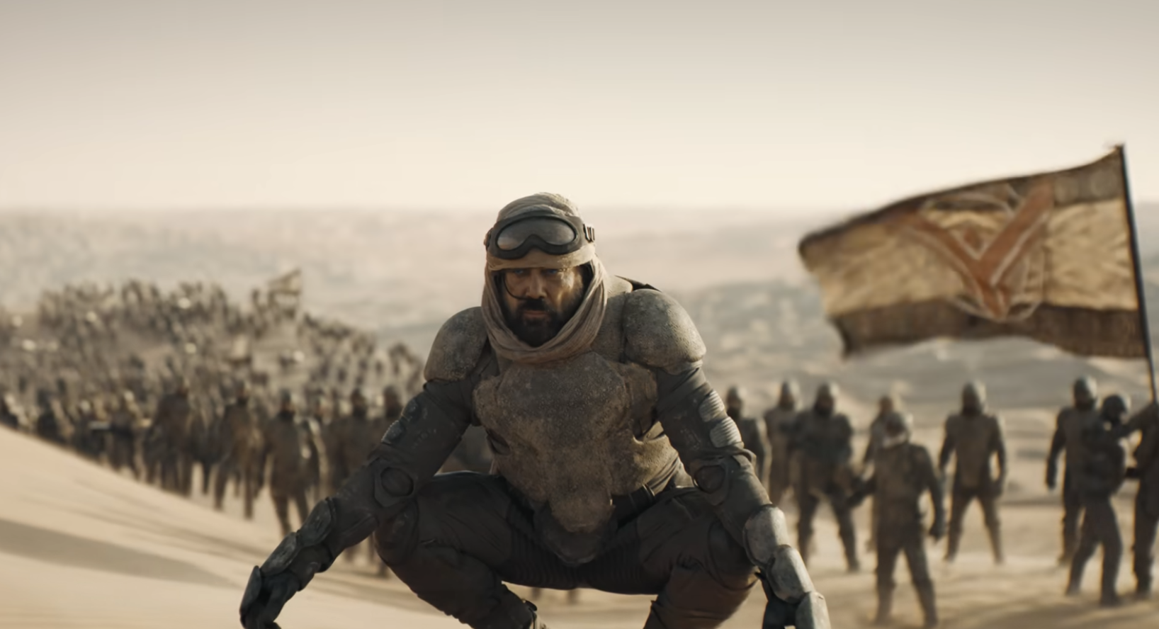 Personaje de película, revestido en armadura de ciencia ficción, posa agachado en un paisaje desértico con gente al fondo