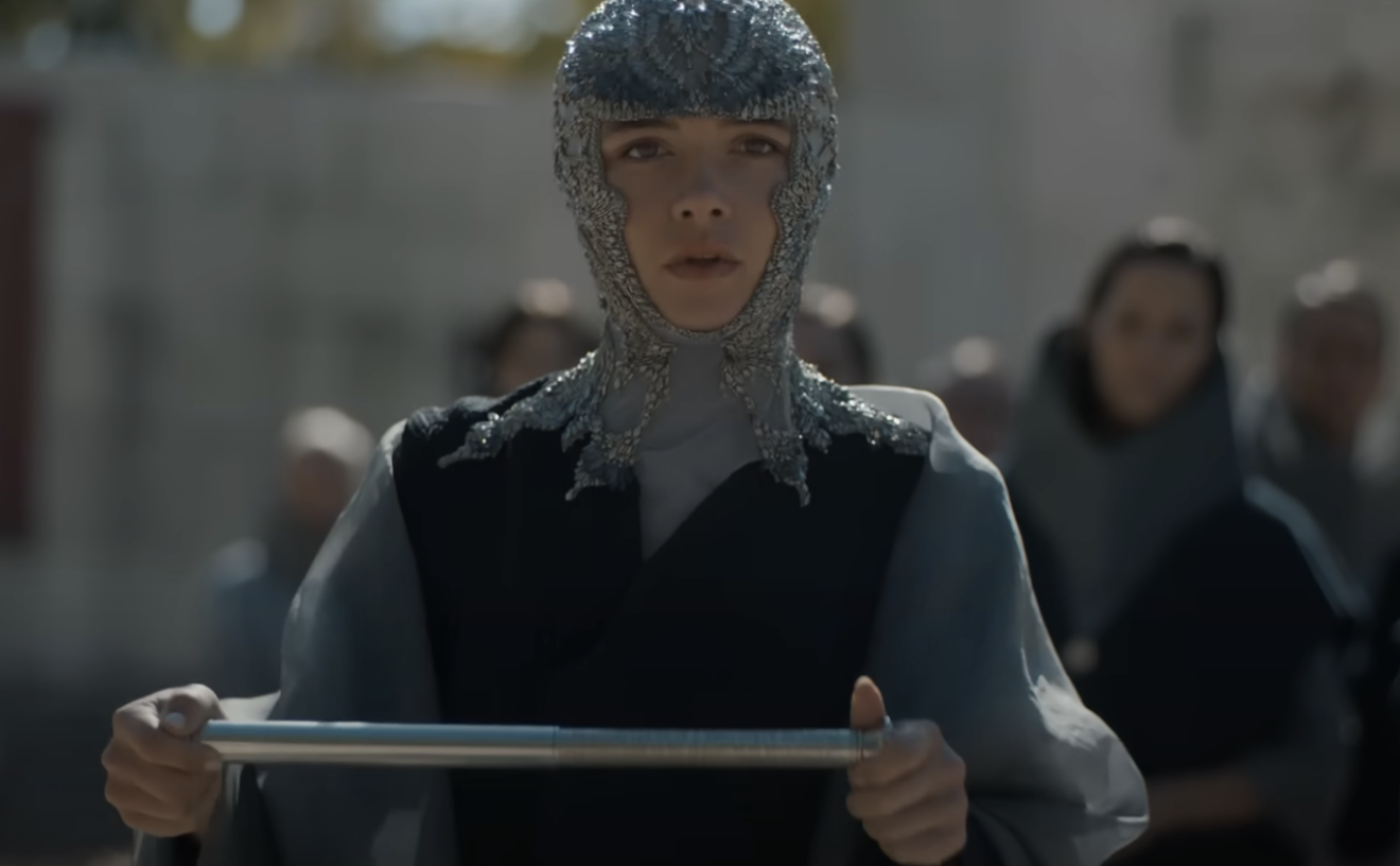 Personaje de ficción con vestimenta medieval y capucha de malla metálica sostiene espada delante de multitud