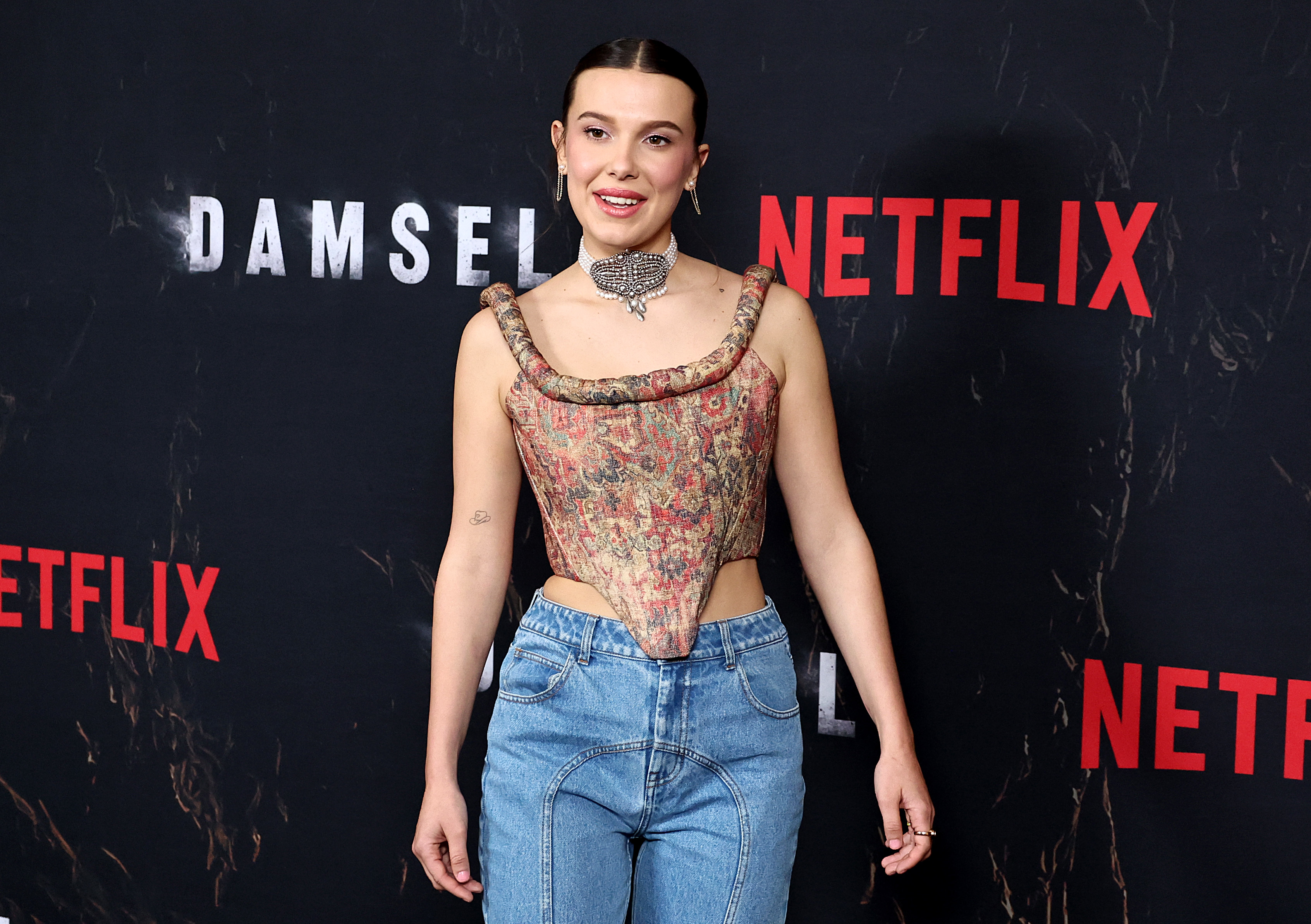 Millie in a patterned corset top and jeans with a statement necklace, posing at a Netflix event