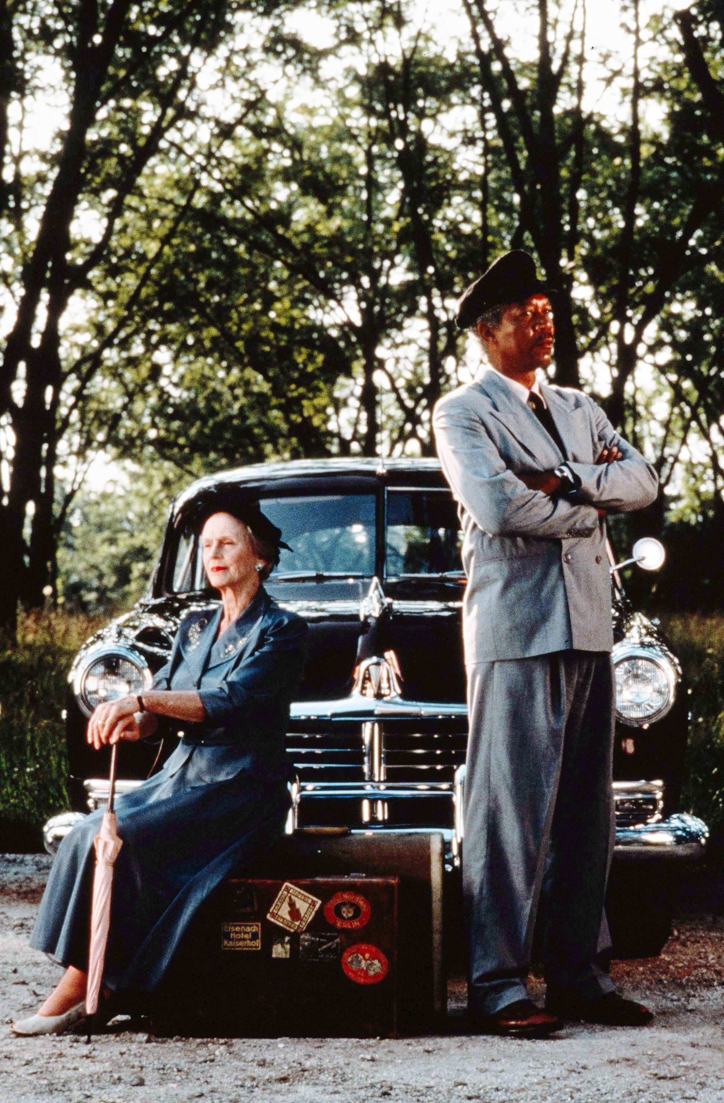 Jessica Tandy as Daisy seated with cane beside standing Morgan Freeman as Hoke Colburn in suit, with a 1940s car behind, in a rural setting