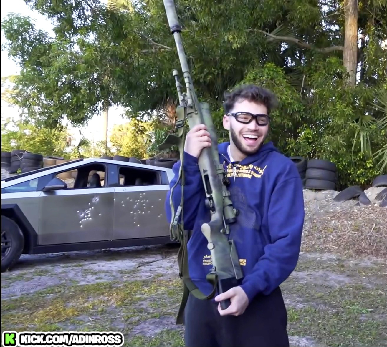 Person holding a large toy gun with a smile, standing outdoors near a car and tires
