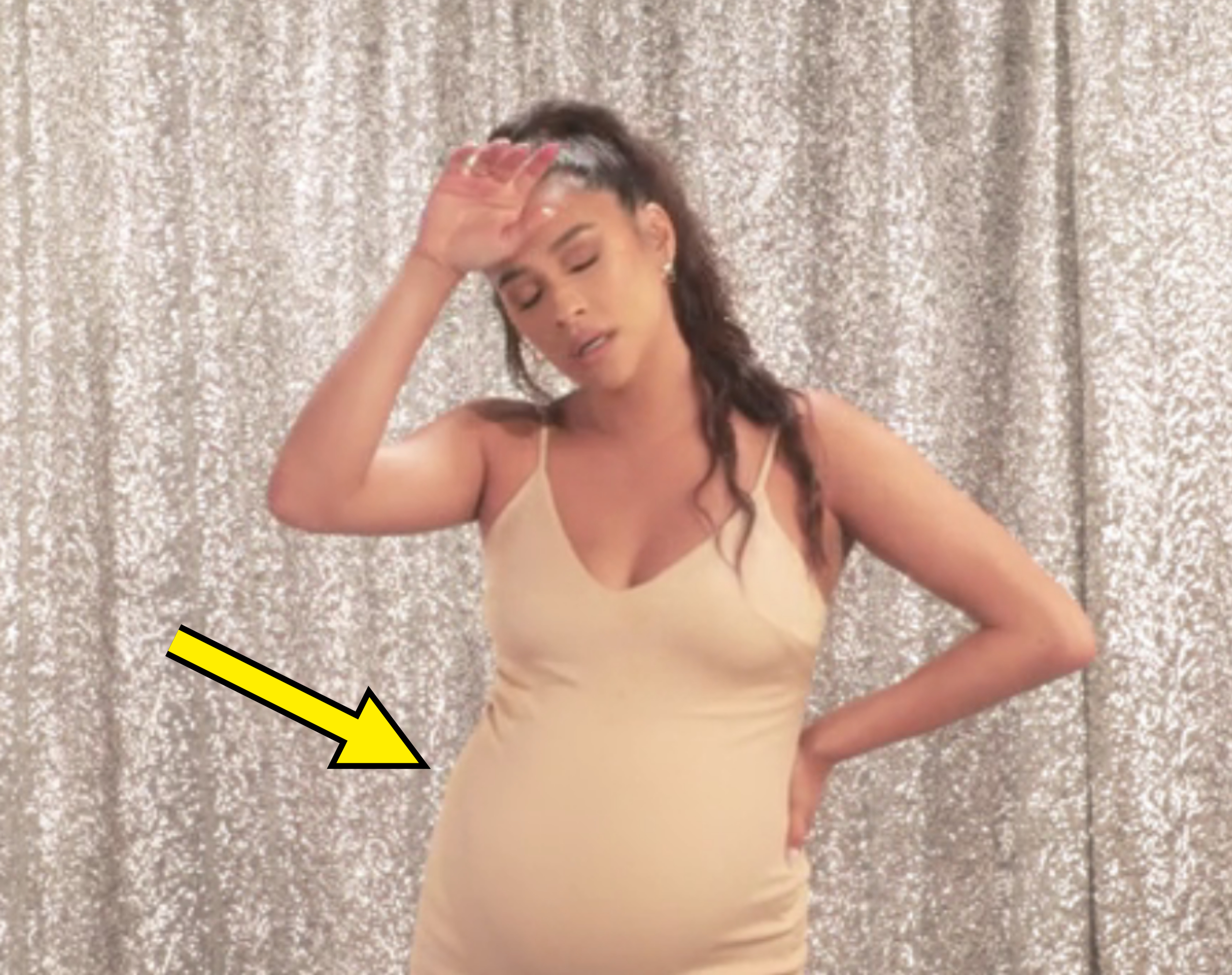 Pregnant woman in a beige dress standing with hand on forehead and hip, appearing tired or in discomfort