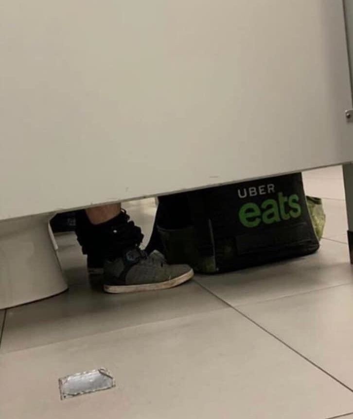 Person sitting in a public restroom stall with an Uber Eats bag visible on the floor under the door