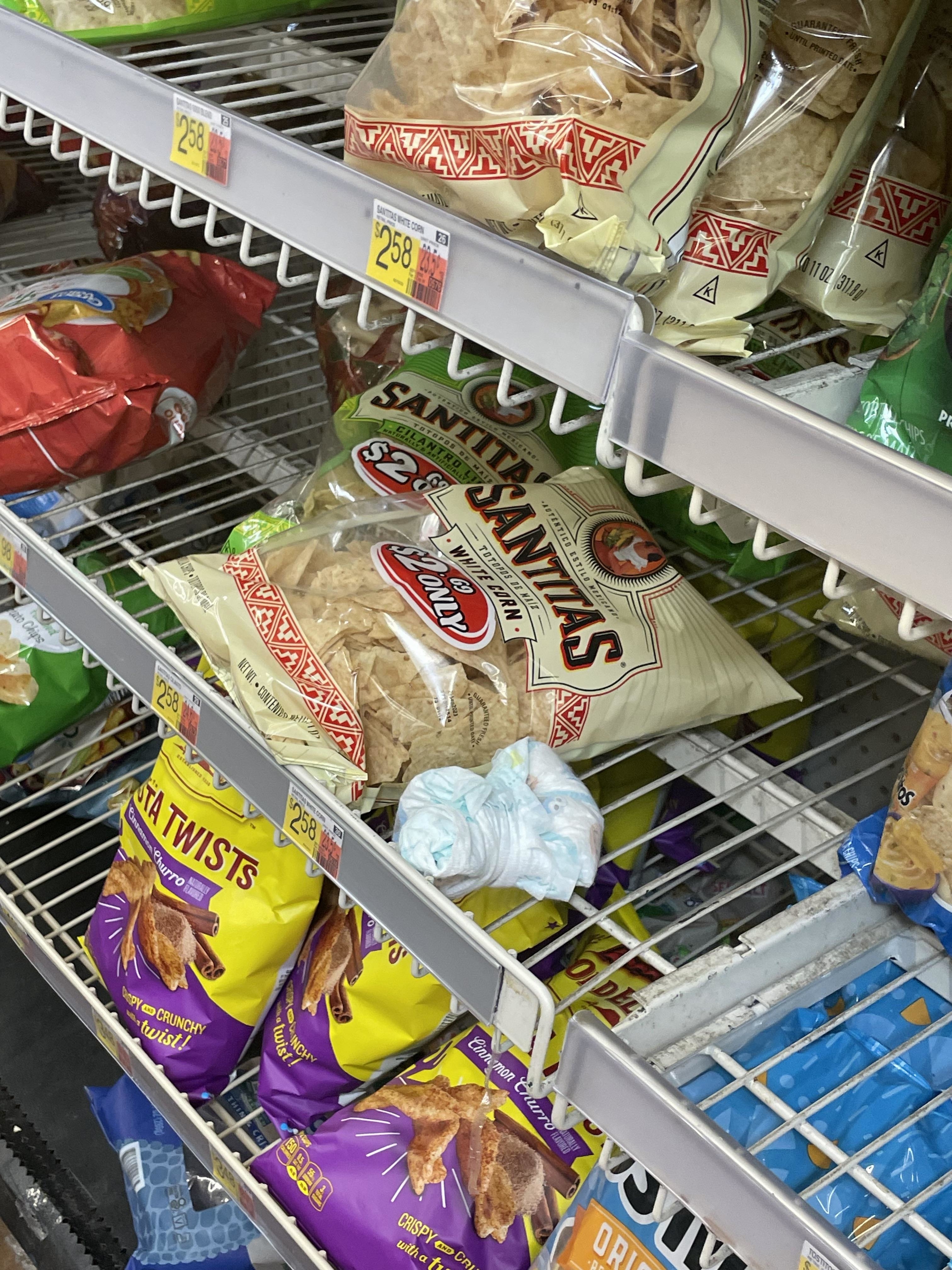 Shelves stocked with various brands of tortilla chips in a store and a dirty diaper on one shelf