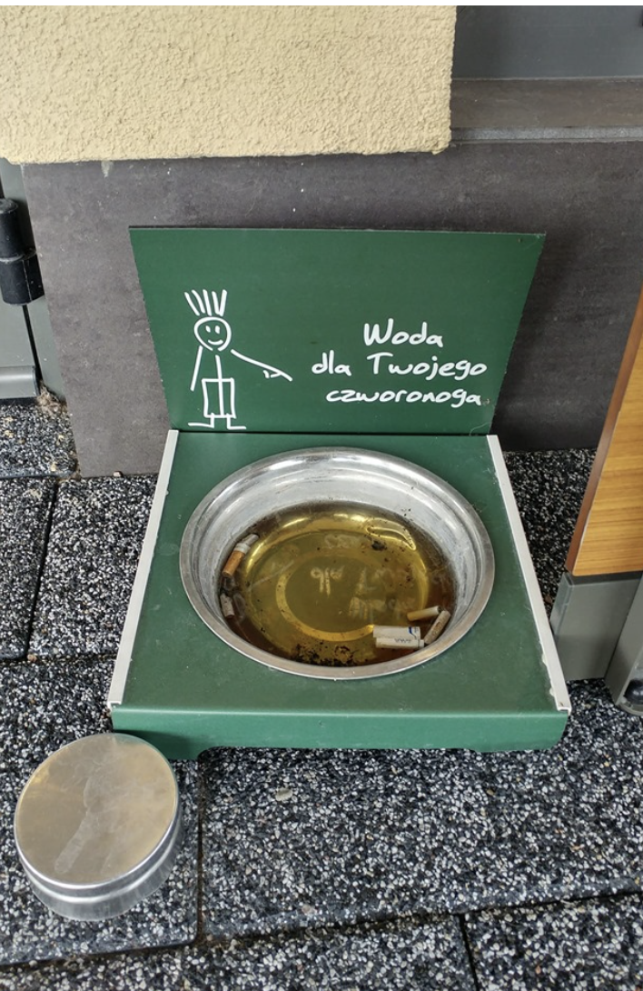 A water station bowl for pets littered with cigarettes and other trash and a sign featuring a pet icon; sign in Polish indicates &quot;water for your pet&quot;