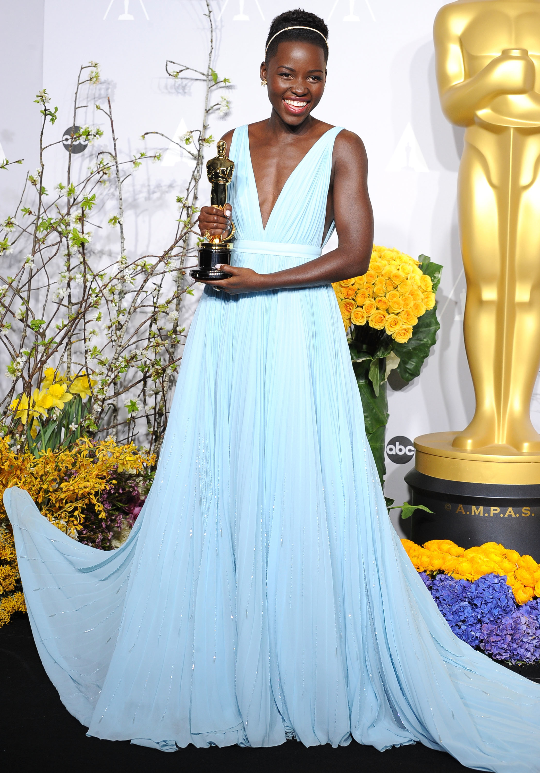 Lupita Nyong&#x27;o holding an Oscar, wearing a plunging V-neck light blue gown with a flowing skirt, standing beside a golden statue