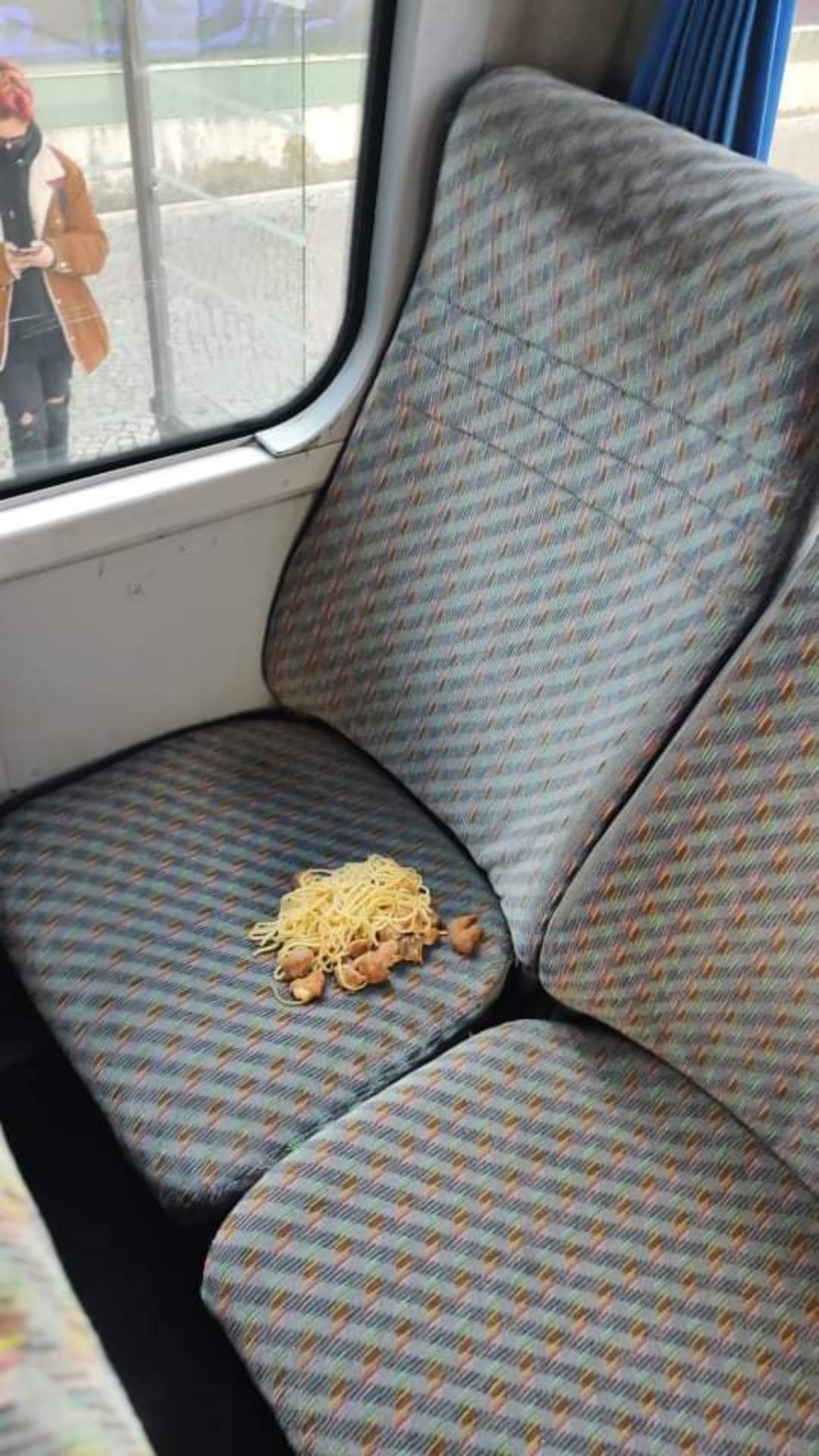 Spaghetti spilled on a patterned seat inside a train, with a passenger standing in the background