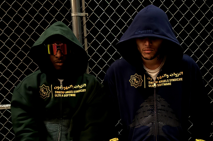 Two individuals in hoodies with text, leaning on a chain-link fence
