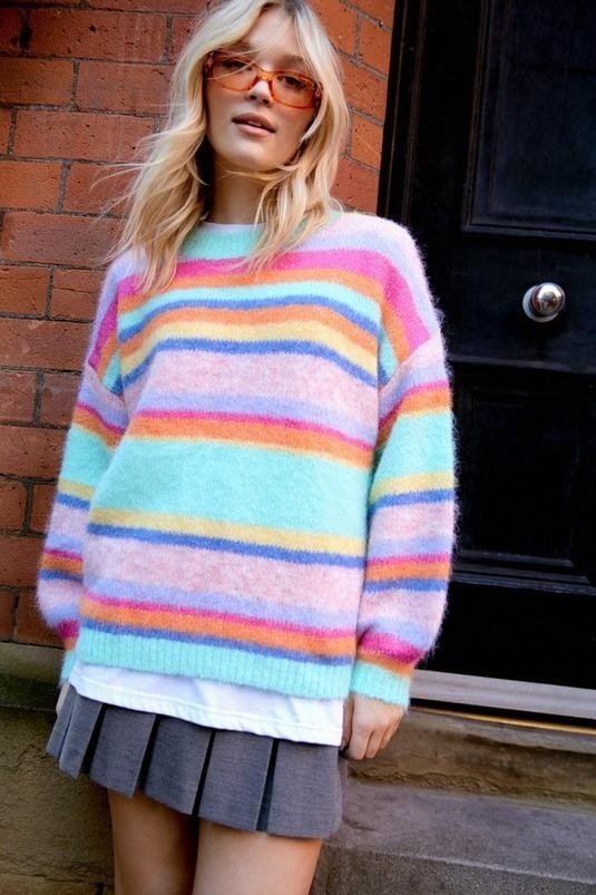 Person in a striped sweater and pleated skirt standing in front of a door