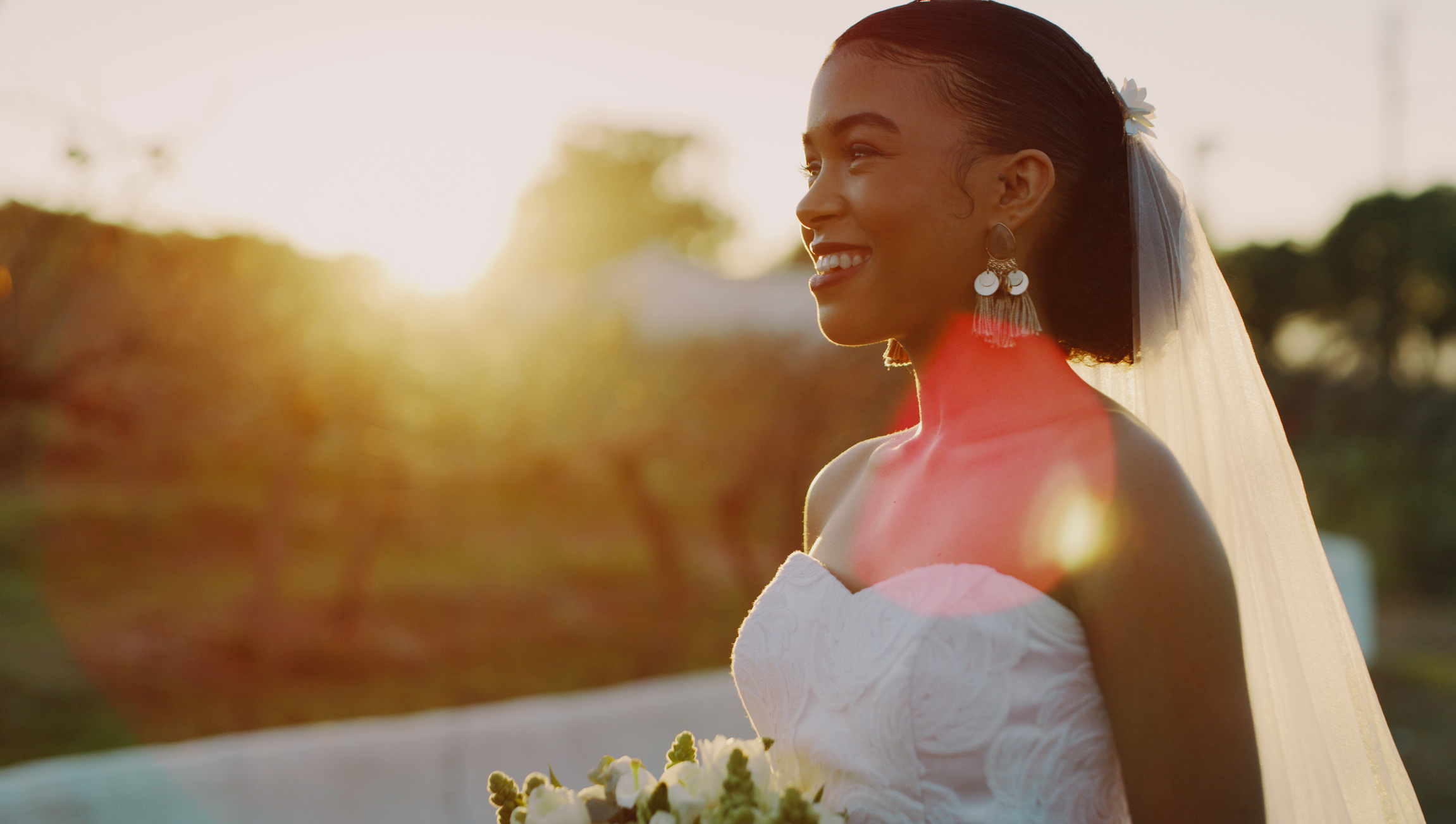 Smiling bride in a strapless wedding dress holding a bouquet, with a sunset background