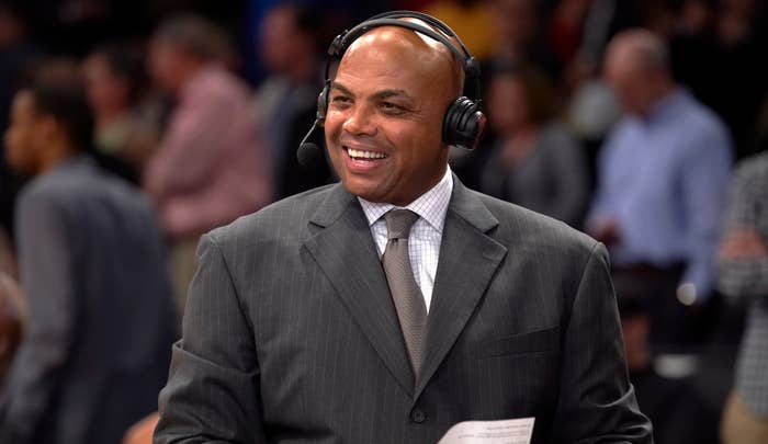 Charles Barkley wearing a suit and headset, smiling while sitting courtside at a basketball game