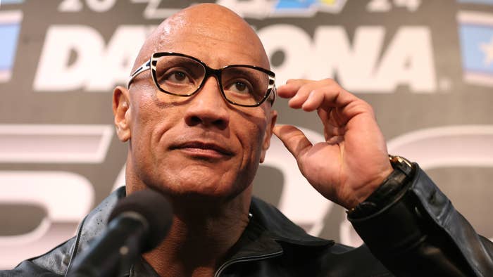 The Rock in formal attire adjusting glasses at a press event