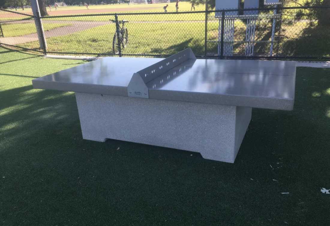 Outdoor ping pong table made of metal and concrete, situated on grass near a fence. No persons in the image