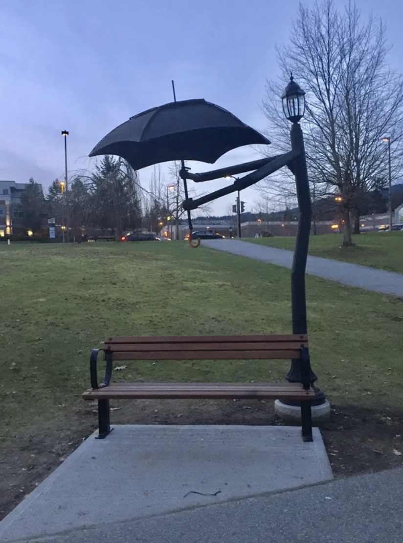 An umbrella hangs above a park bench, attached to a lamp post, in an outdoor setting at dusk