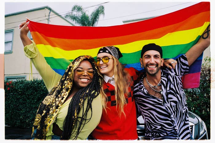 Three friends smiling with a Pride flag. They wear stylish casual outfits