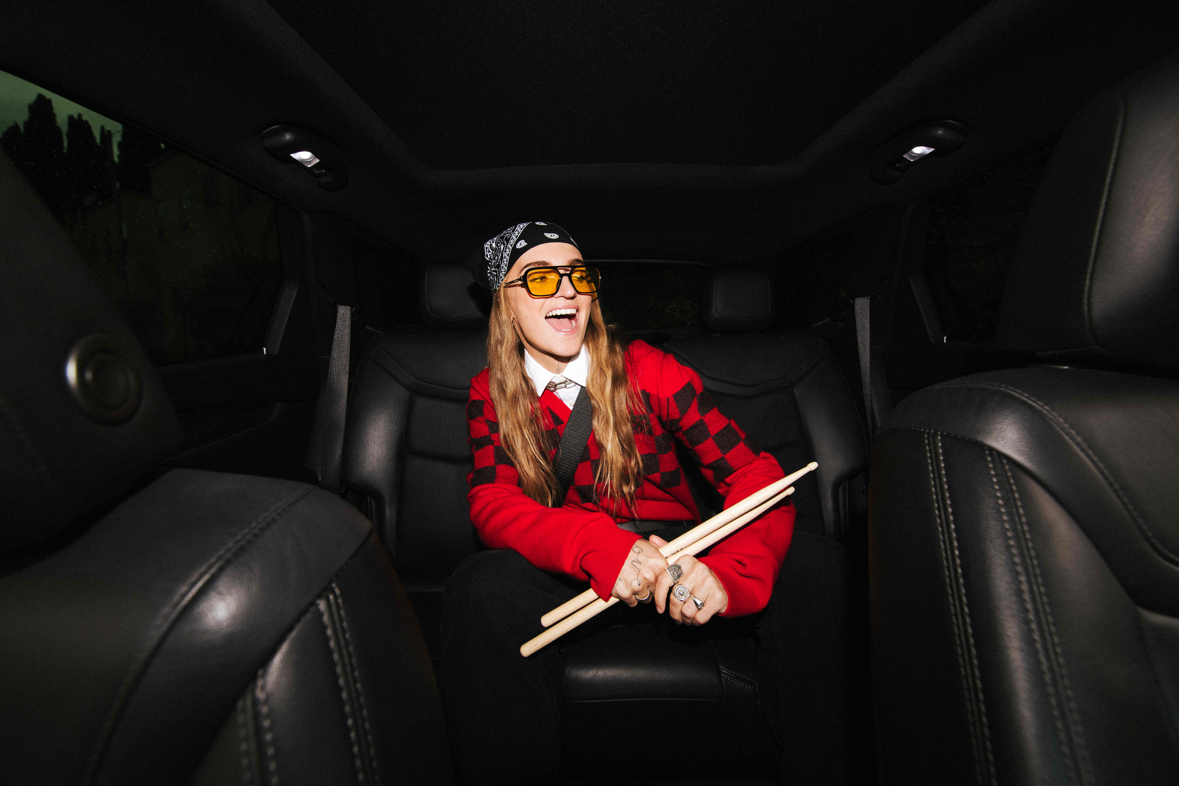 Person in school-inspired outfit with tie and sweater, holding drumsticks, seated in a car