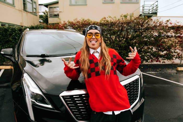Individual in a checkered sweater and cap stands in front of a car making a peace sign with both hands