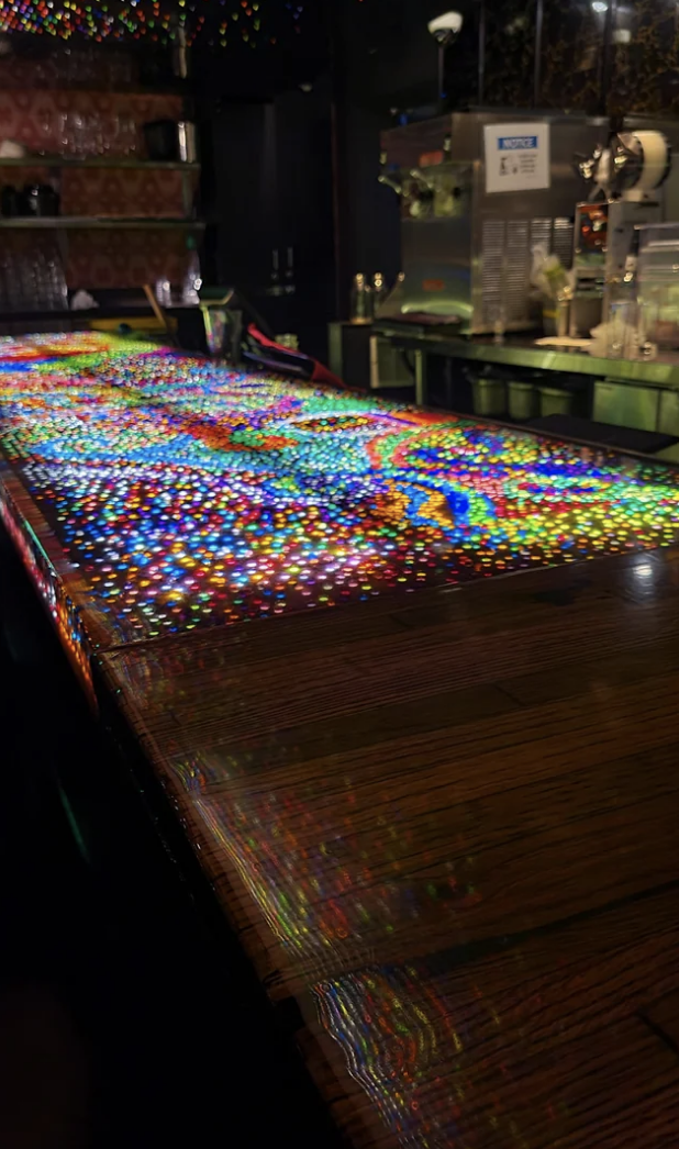 A bar counter with an intricate, multicolored light display, casting vibrant reflections. No people in the image