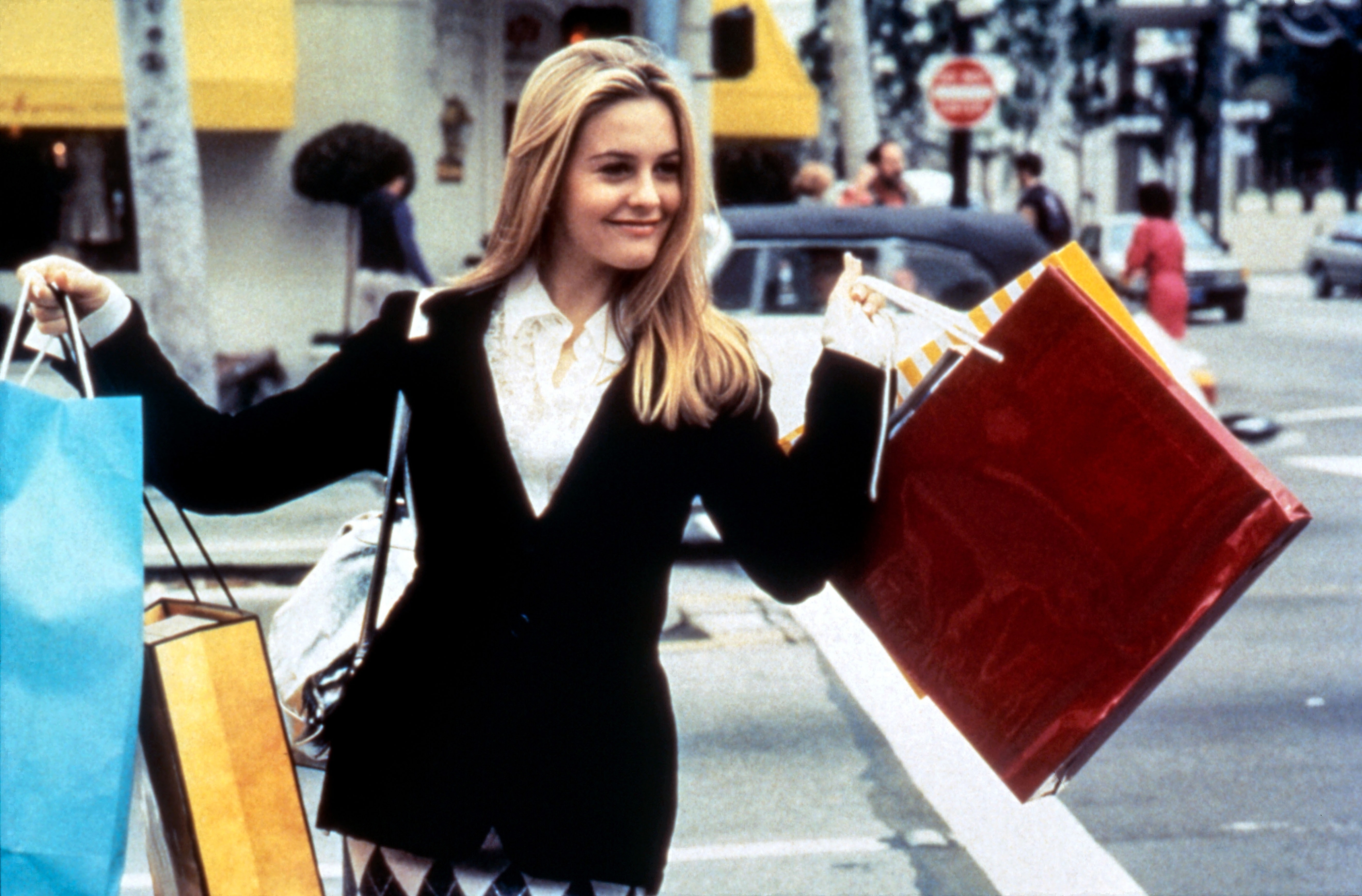 Cher Horowitz from Clueless smiling, holding shopping bags on a city street. Wearing a blazer and skirt
