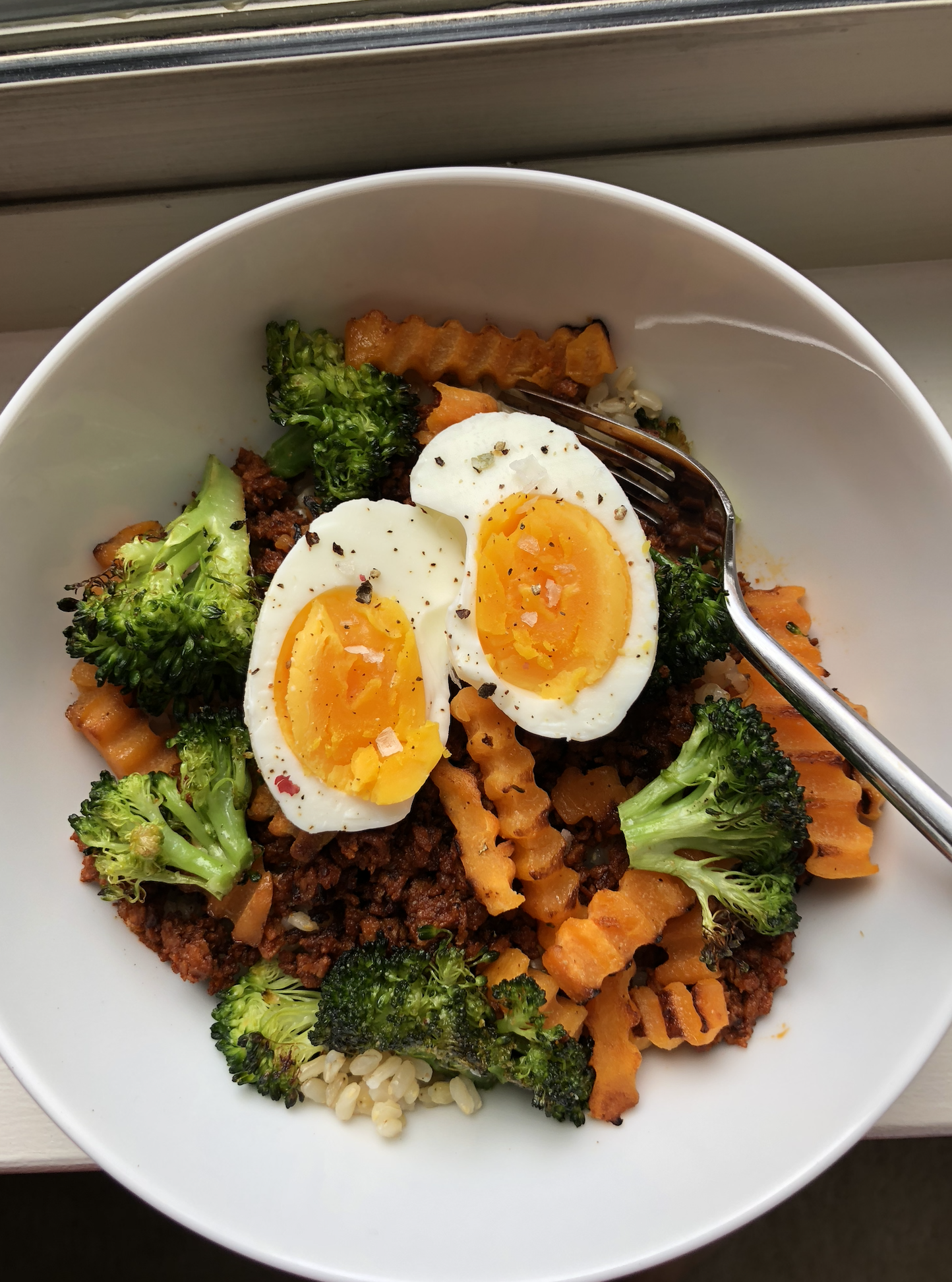 Bowl of food with quinoa, roasted broccoli, wavy-cut sweet potatoes, topped with a soft-boiled egg