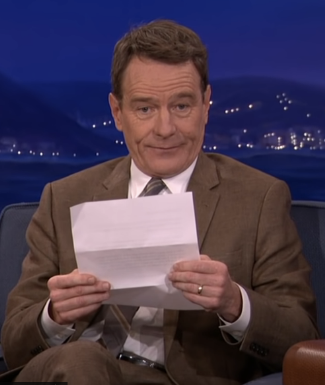 Bryan Cranston looks uncomfortable while holding the note in his hand, seated during a talk show