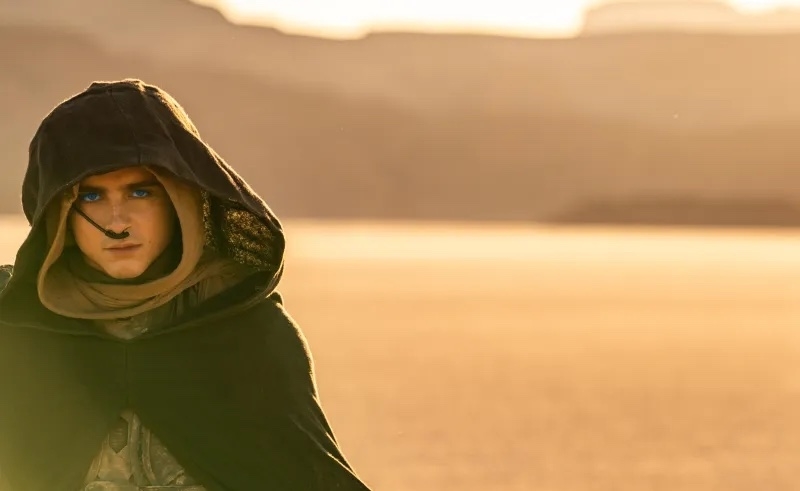 Person in a hooded cloak with intense gaze, in a desert setting, evoking a cinematic scene