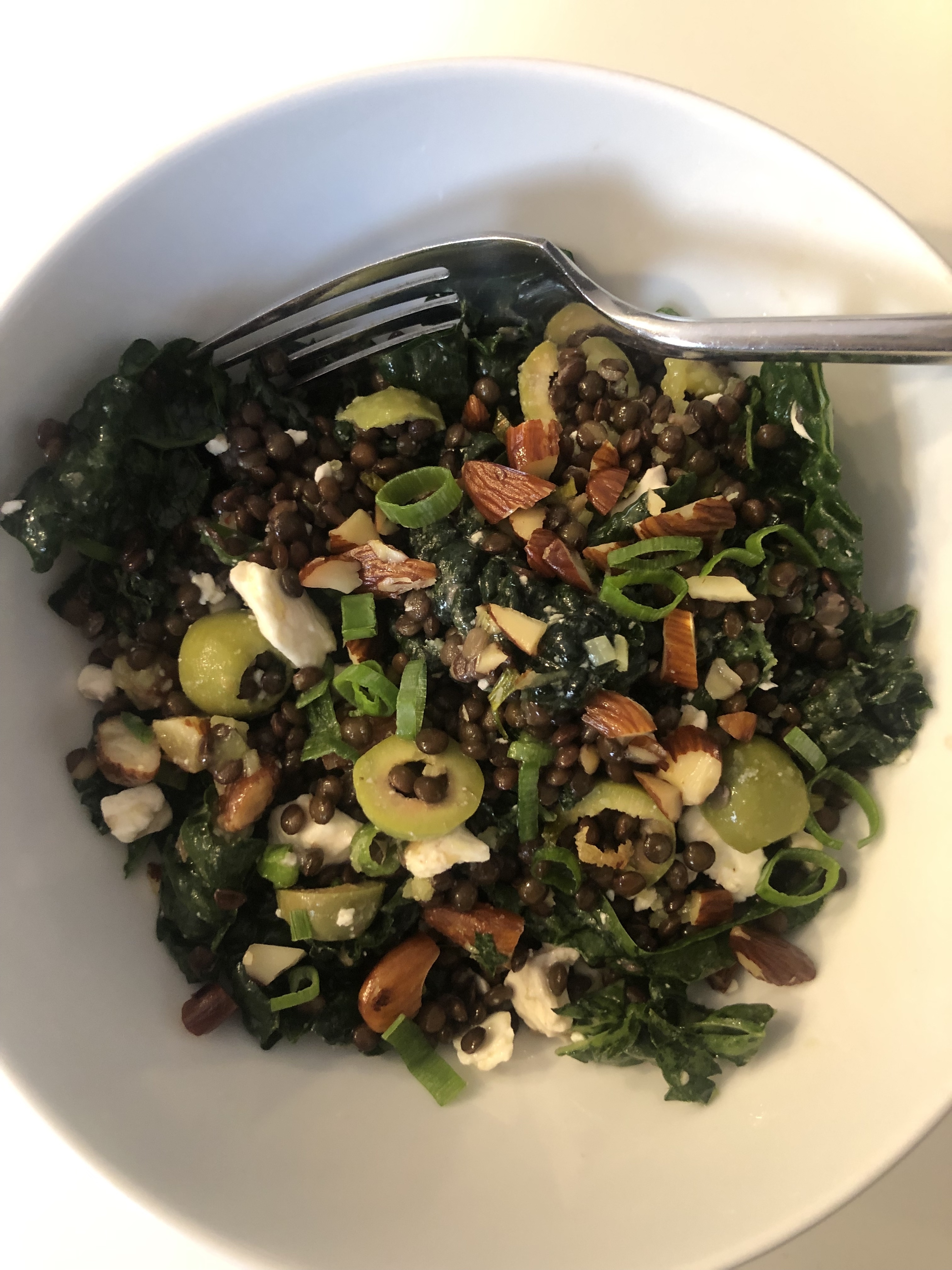 A bowl of lentil salad with kale, nuts, and sliced vegetables, with a fork