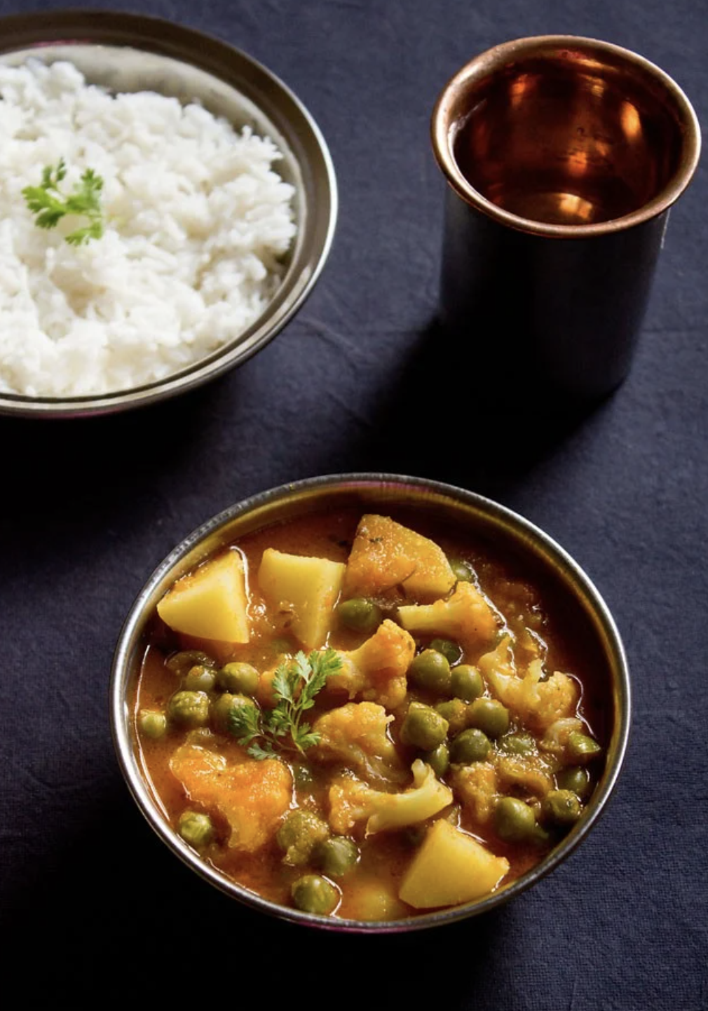 Aloo matar (potato and pea curry) served with rice and a cup, on a dark surface